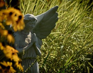 Praying angel stands amongst the flowers in the garden on a sunny day in spring.