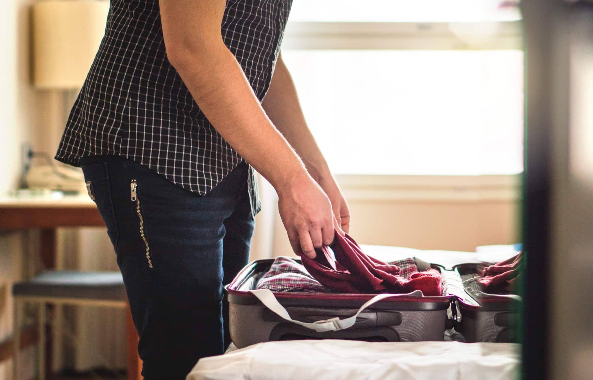 Packing suitcase in hotel room. Young man folding t-shirt on baggage in home bedroom. Open luggage on bed for vacation preparing.