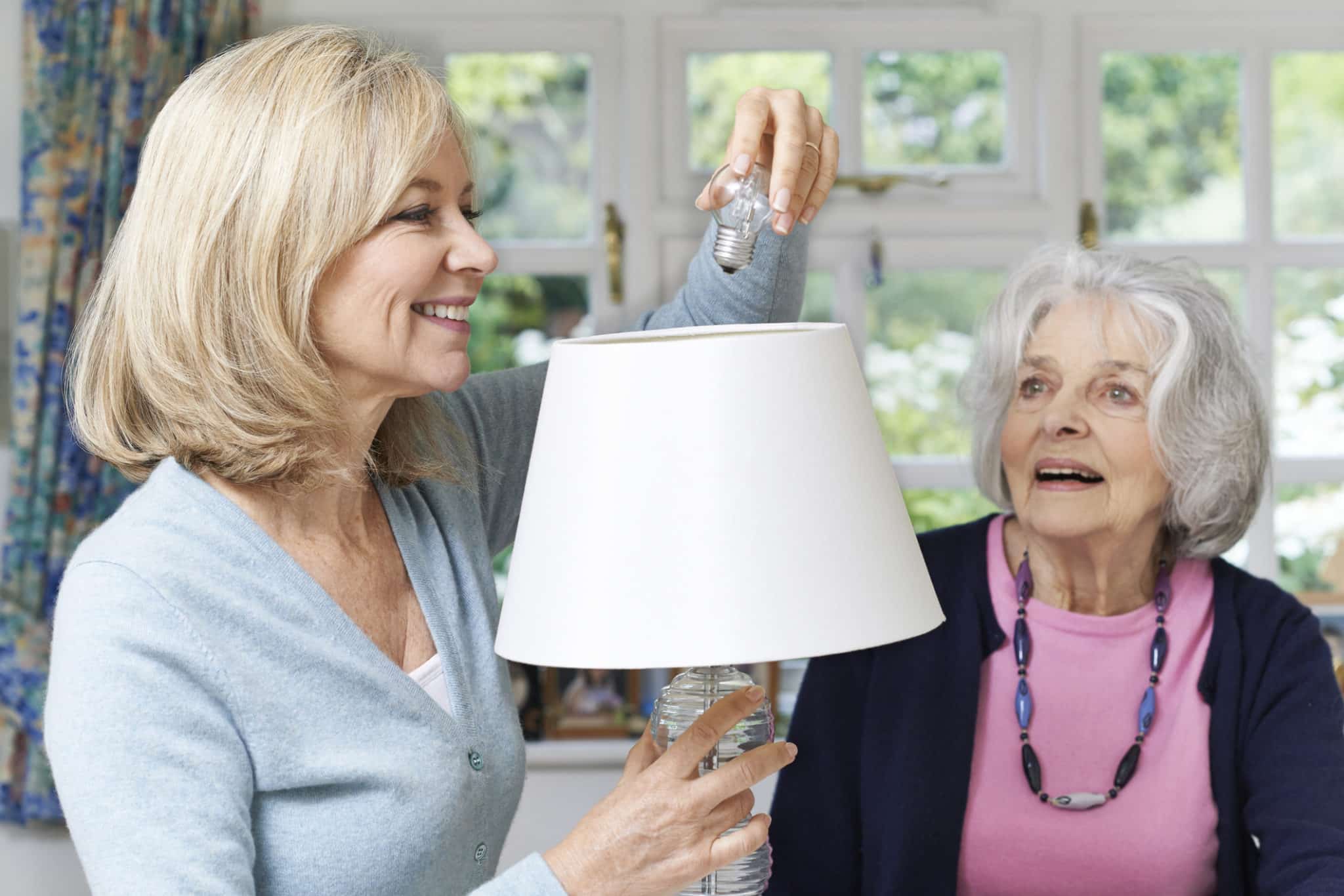 Female Neighbor Helping Senior Woman Change Lightbulb In Lamp