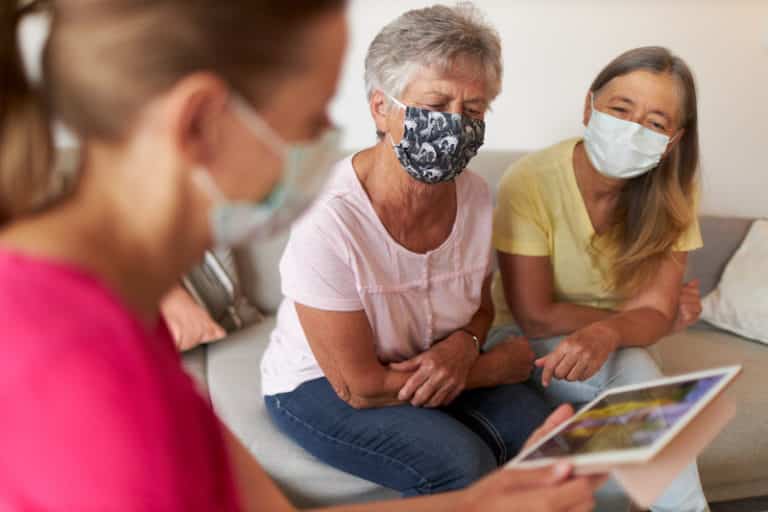 A grandmother sits with her daughter and granddaughter on the sofa in the living room and uses the Ipad to make calls while everyone has to wear a protective mask or surgical mask