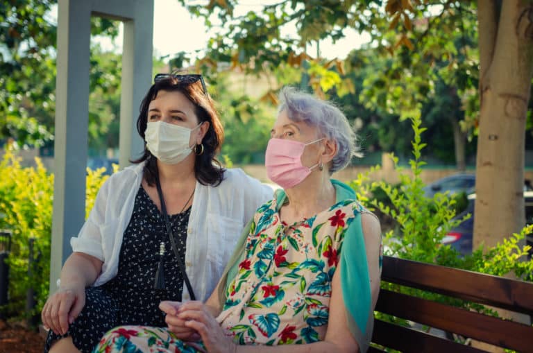 Caregiver and the old woman, wearing masks, sat down to rest on a park bench near the house.