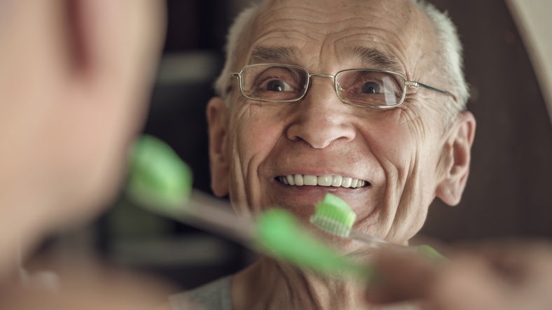 Gray haired senior man is cleaning his teeth and smiling looking in mirror